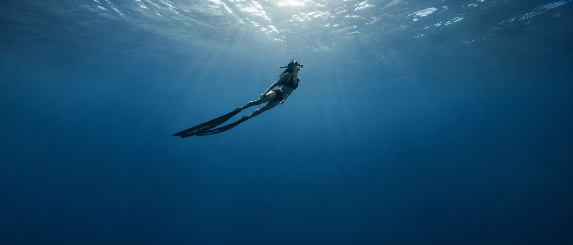 A freediver glides through deep blue water under rays of sunlight.